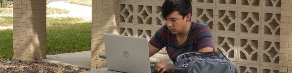 male student sitting at a picnic table typing on silver laptop