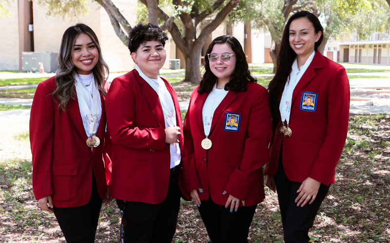 Four Cosmetology students pose at the Uvalde Campus for a photo wearing matching red jackets and medals earned during a SkillsUSA state competition earlier this year.