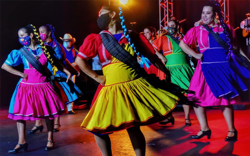 A group of Guadalupe Dance Company members perform on a stage in bright, colorful dresses.